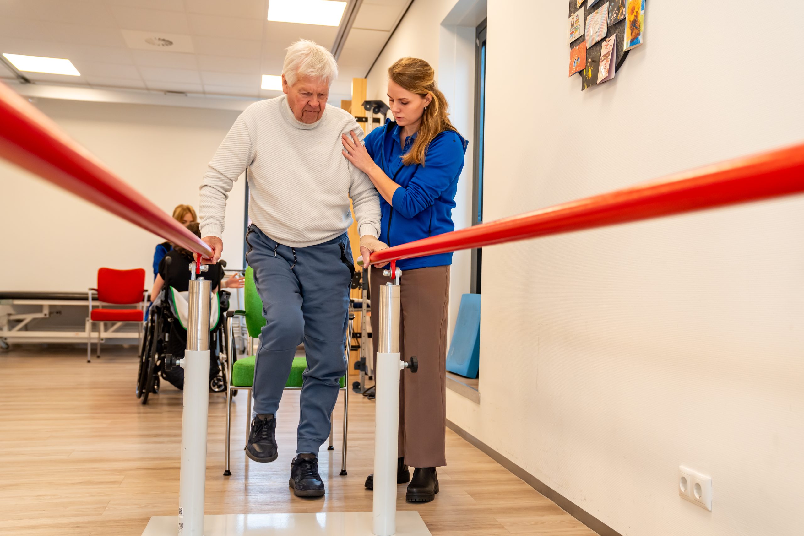 Oudere man oefent lopen tussen parallelle stangen met hulp van fysiotherapeut.