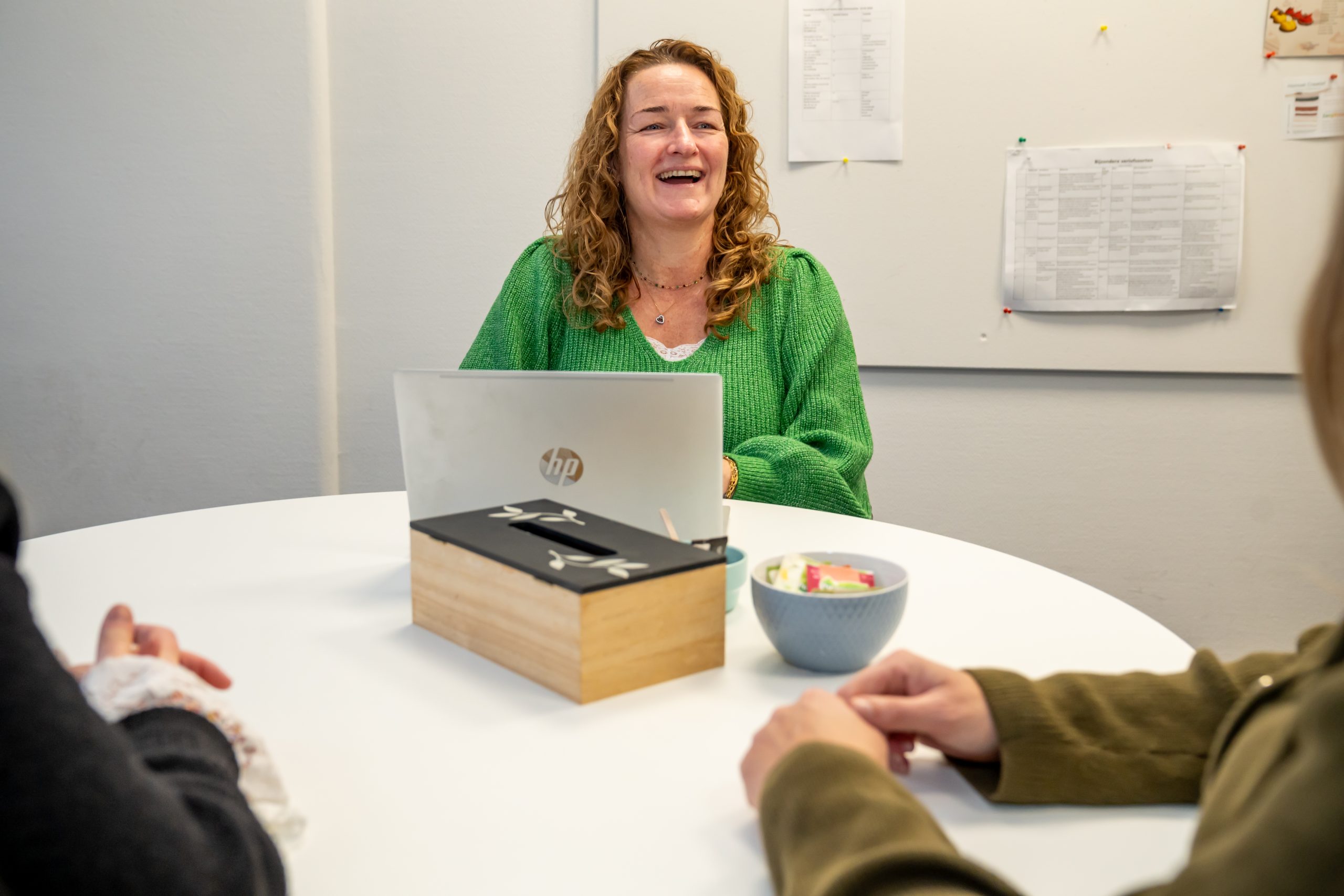 Vrouw in groene trui lacht tijdens overleg aan ronde tafel met laptop.