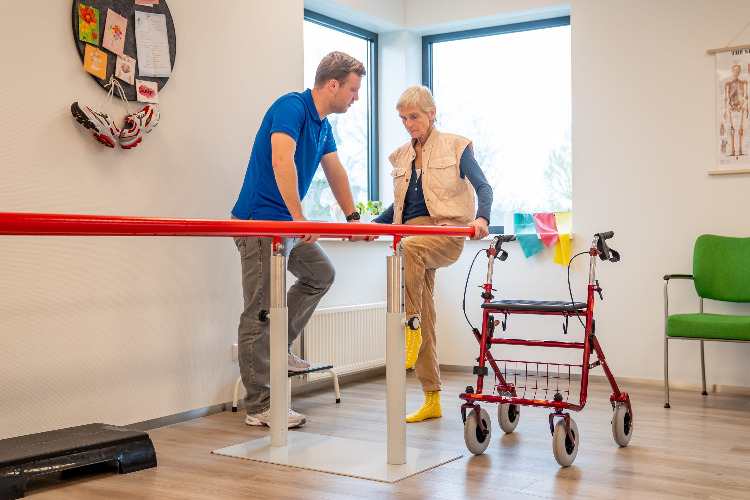 Fysiotherapeut begeleidt oudere vrouw bij loopoefeningen aan parallelle oefenbalken, met rollator erbij.