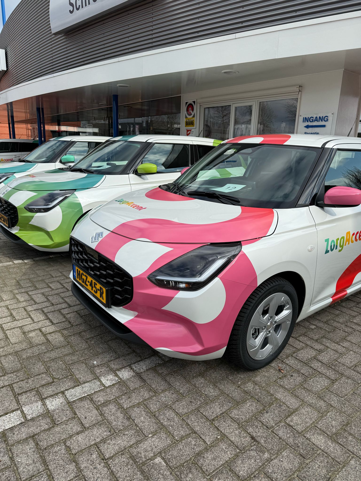 Drie felgekleurde bestickerde bedrijfsauto's geparkeerd voor een pand met bord 'INGANG'.