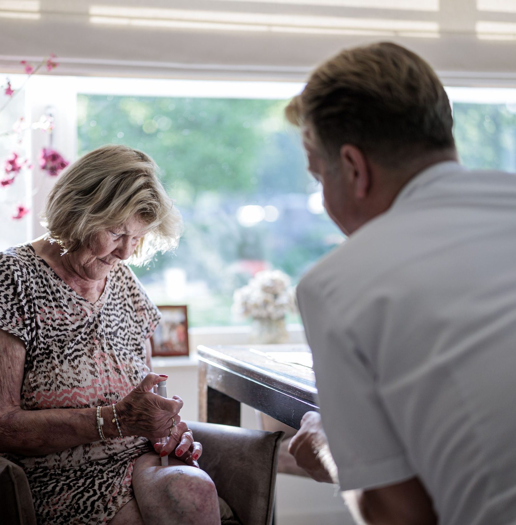 Oudere vrouw spuit medicatie in haar dij, begeleid door zorgverlener in de woonkamer.