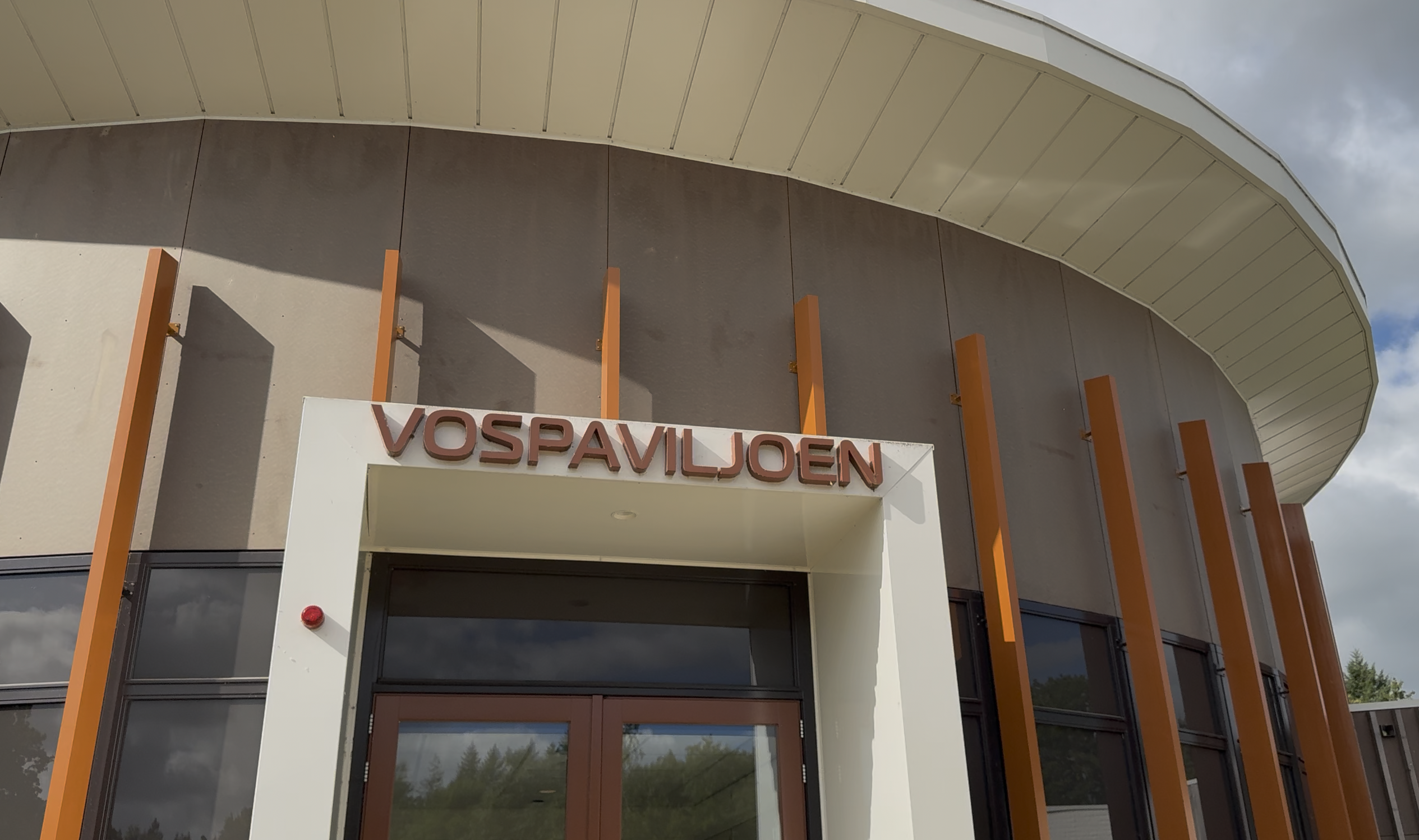 Ingang van het VosPaviljoen met oranje lamellen en ronde gevel onder bewolkte lucht.