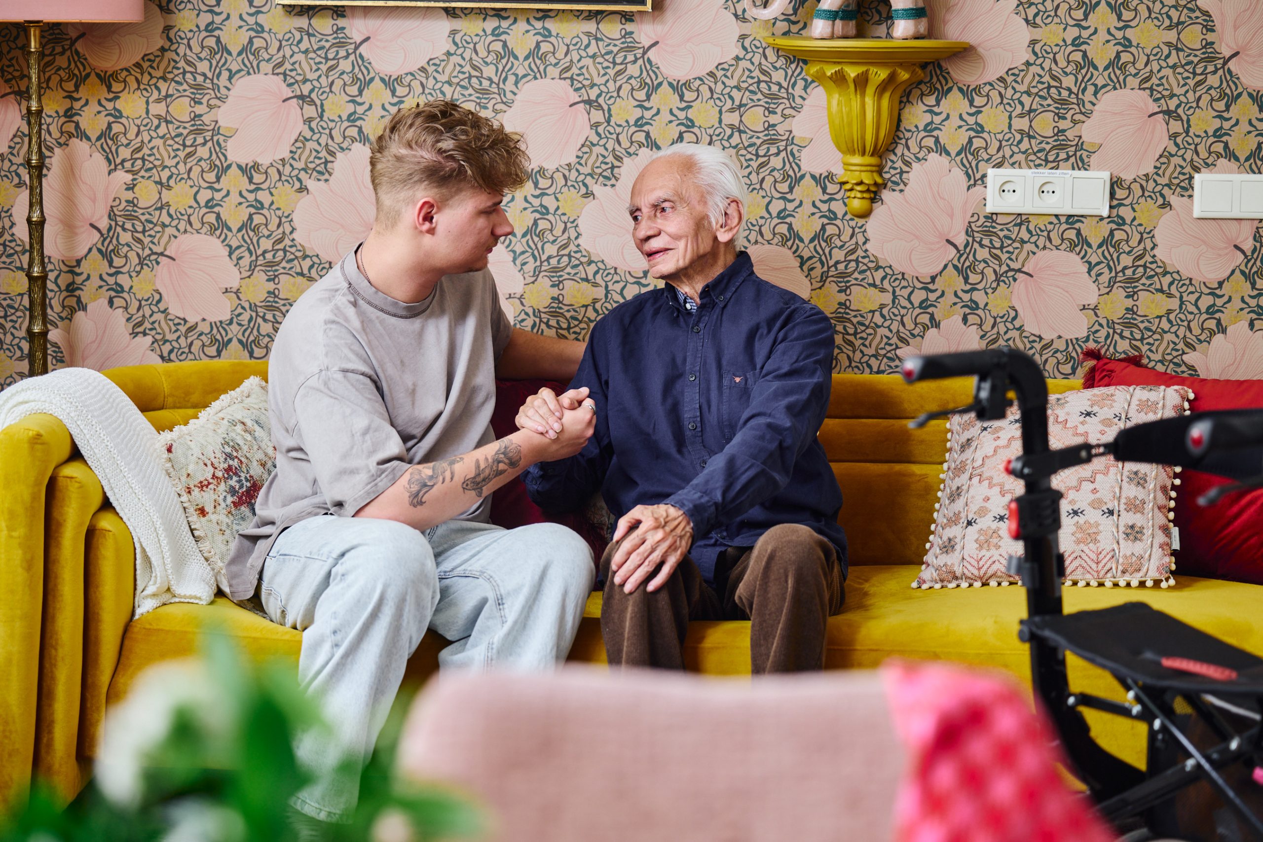 Jonge man en oudere man op gele bank, houden elkaars hand vast in kleurrijke woonkamer met rollator.