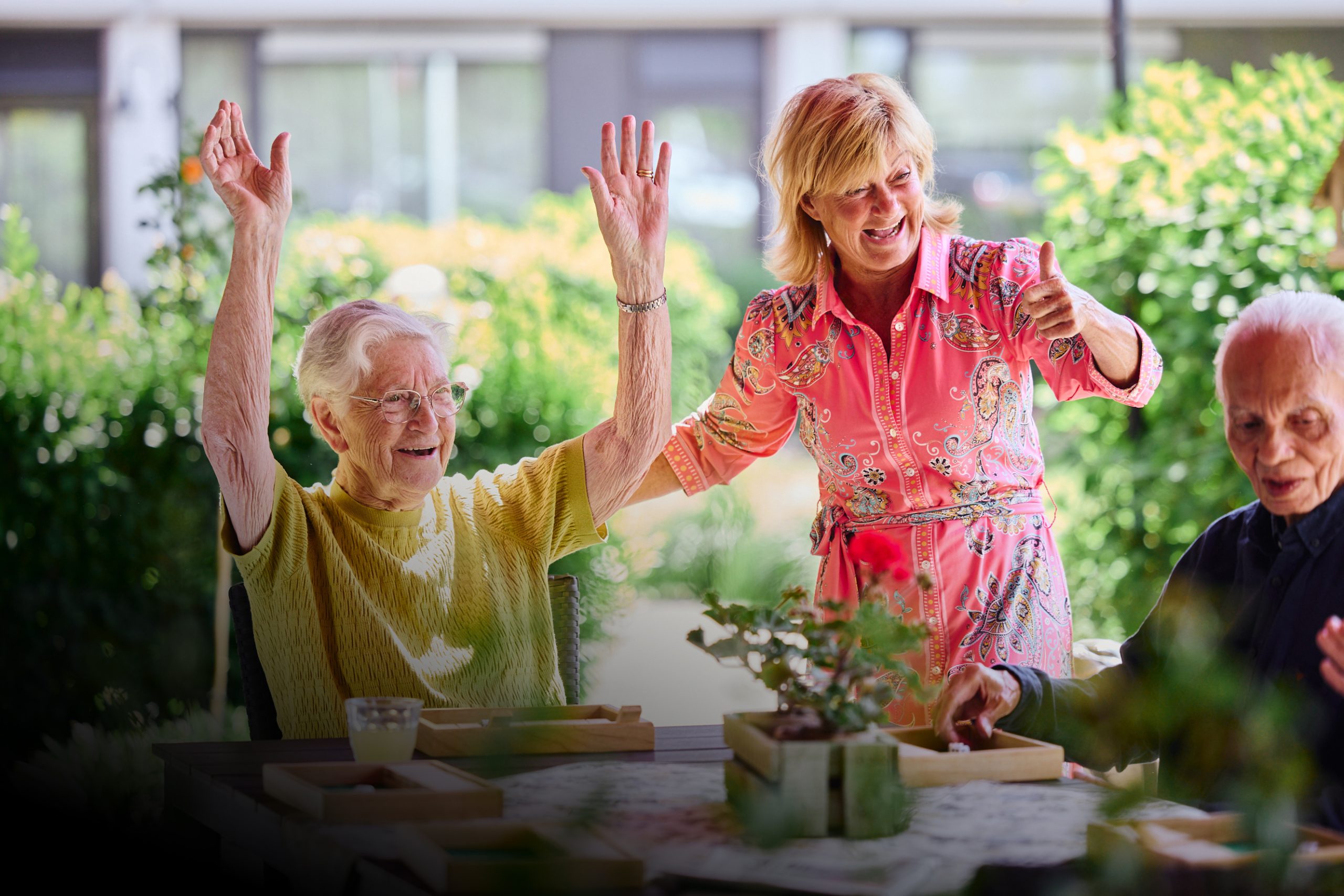 Oudere vrouw juicht met armen omhoog; begeleidster glimlacht en steekt duim op, man speelt aan buitentafel.
