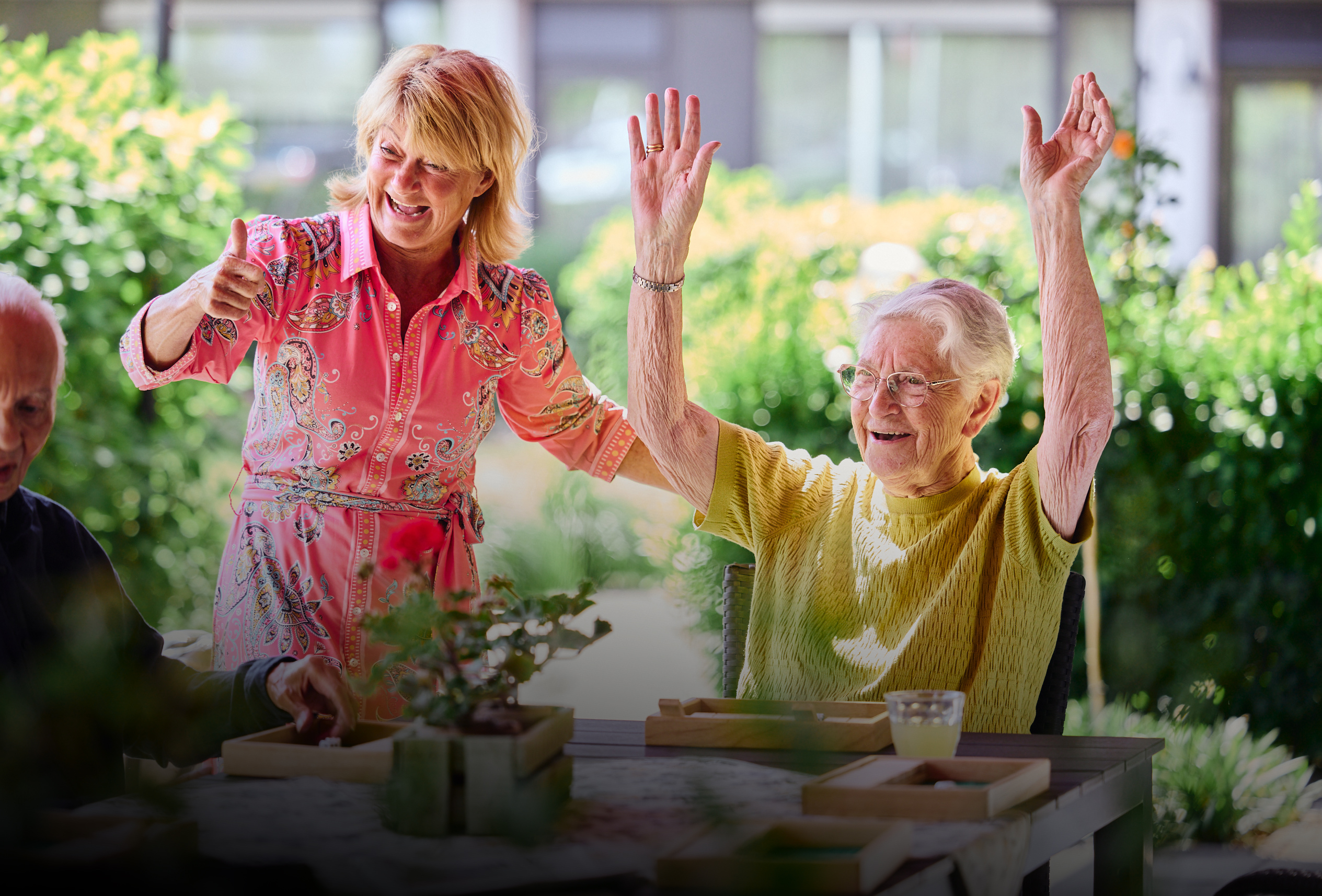 Blije oudere vrouw steekt juichend haar armen omhoog; begeleider naast haar duimt omhoog buiten.