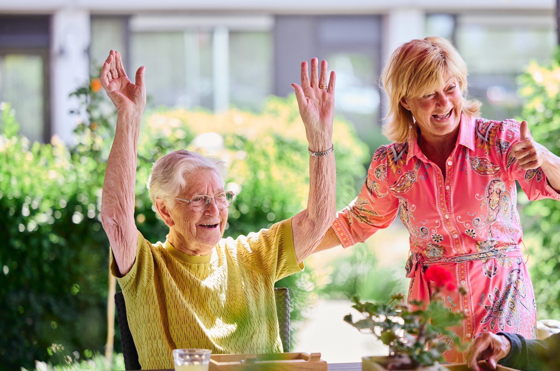 Blije oudere vrouw heft haar armen omhoog; vrouw in roze jurk geeft duim omhoog in zonnige tuin.