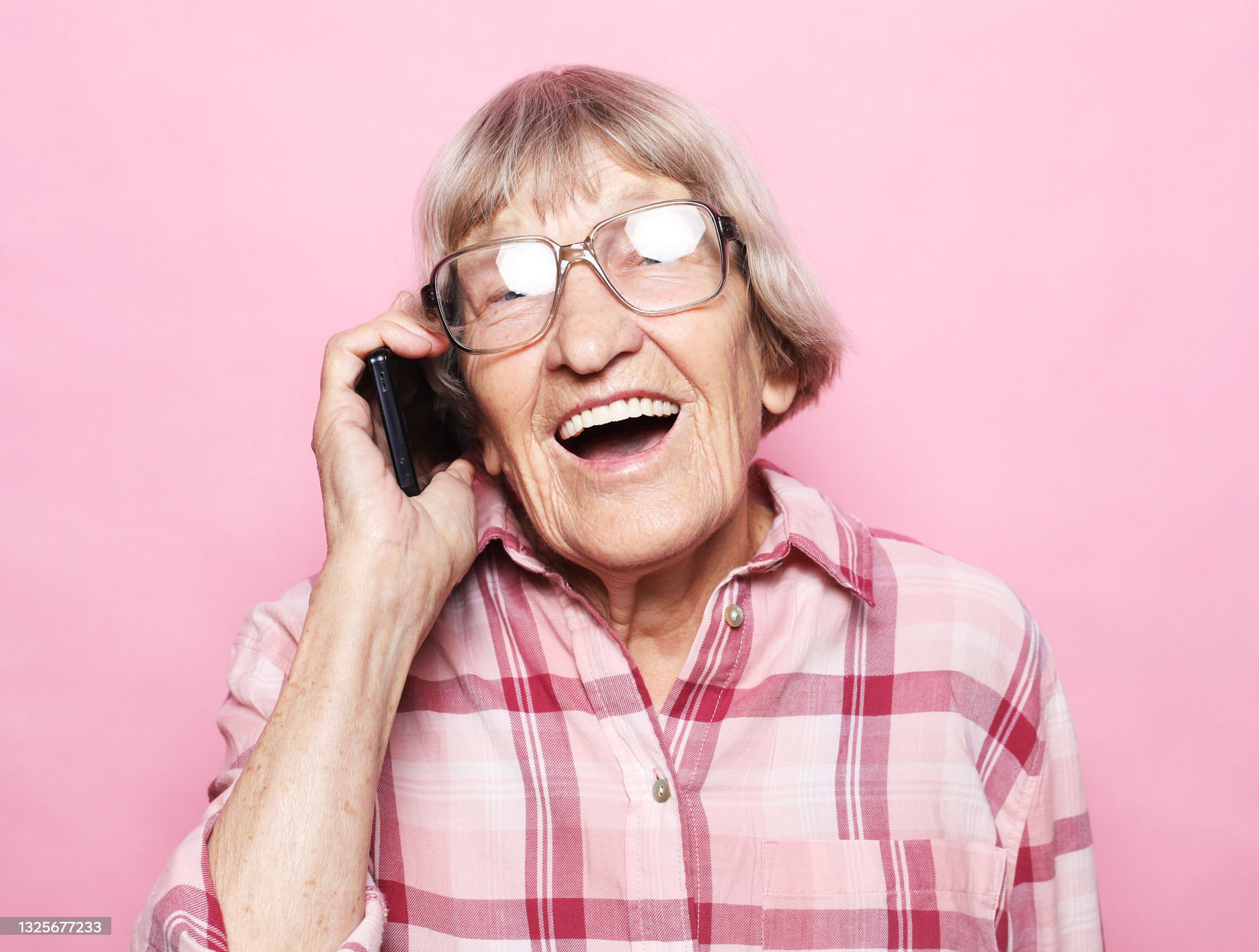 Lachende oudere vrouw met bril belt met smartphone, in roze ruitjesoverhemd voor roze achtergrond.