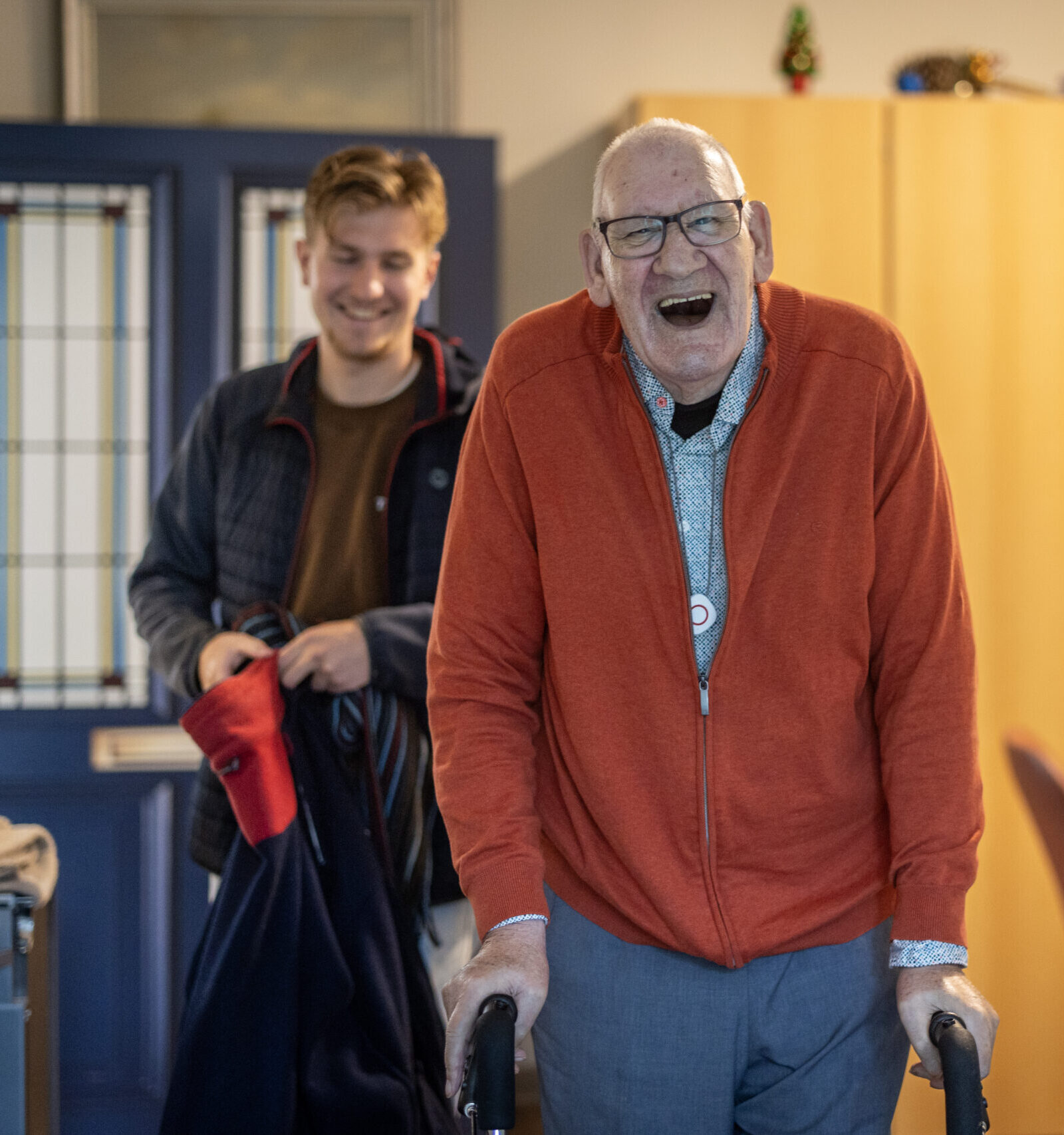 Lachende oudere man met rollator binnen; jonge man staat op de achtergrond.
