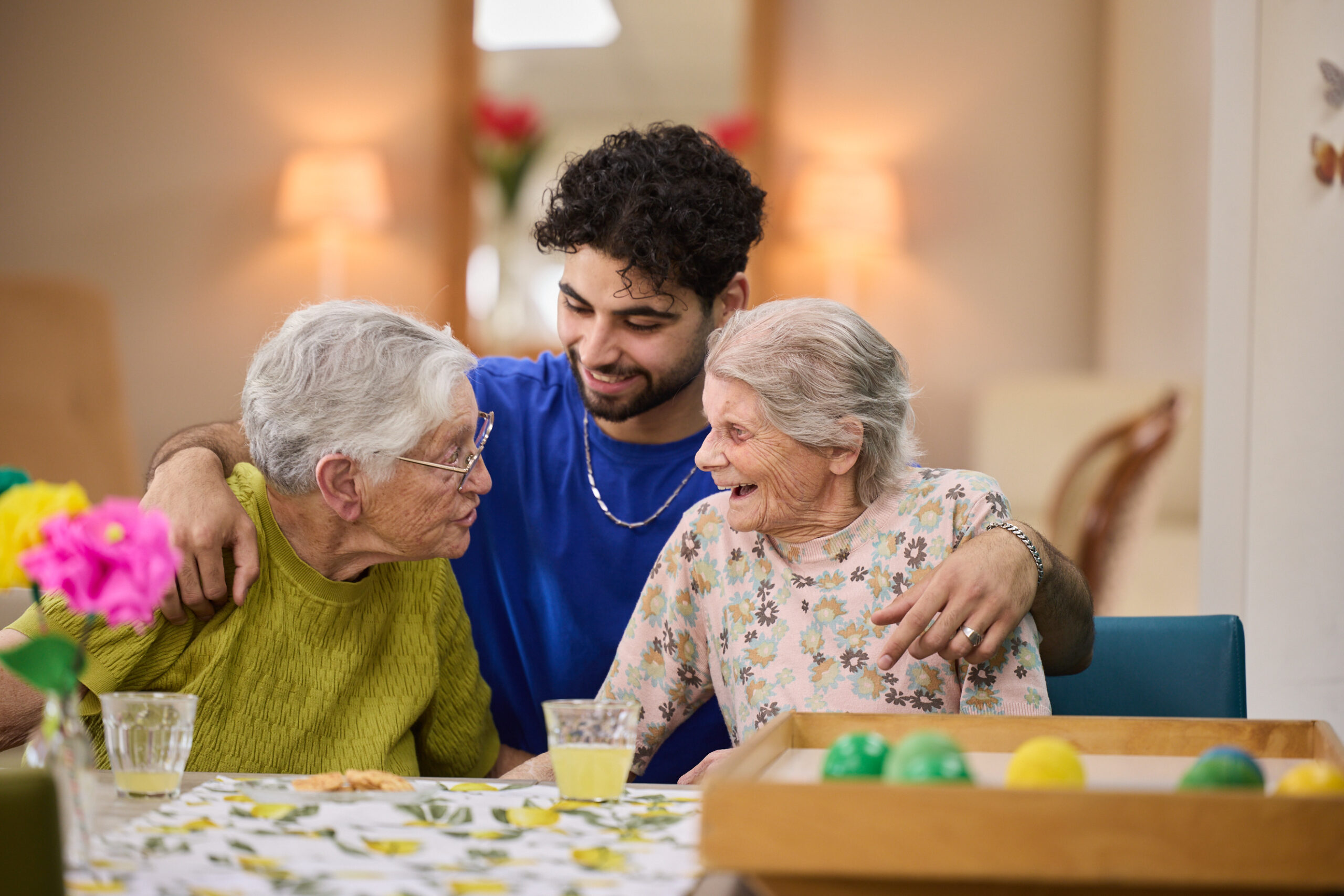 Jonge begeleider omarmt twee lachende oudere vrouwen aan tafel, met limonade en spelballen.