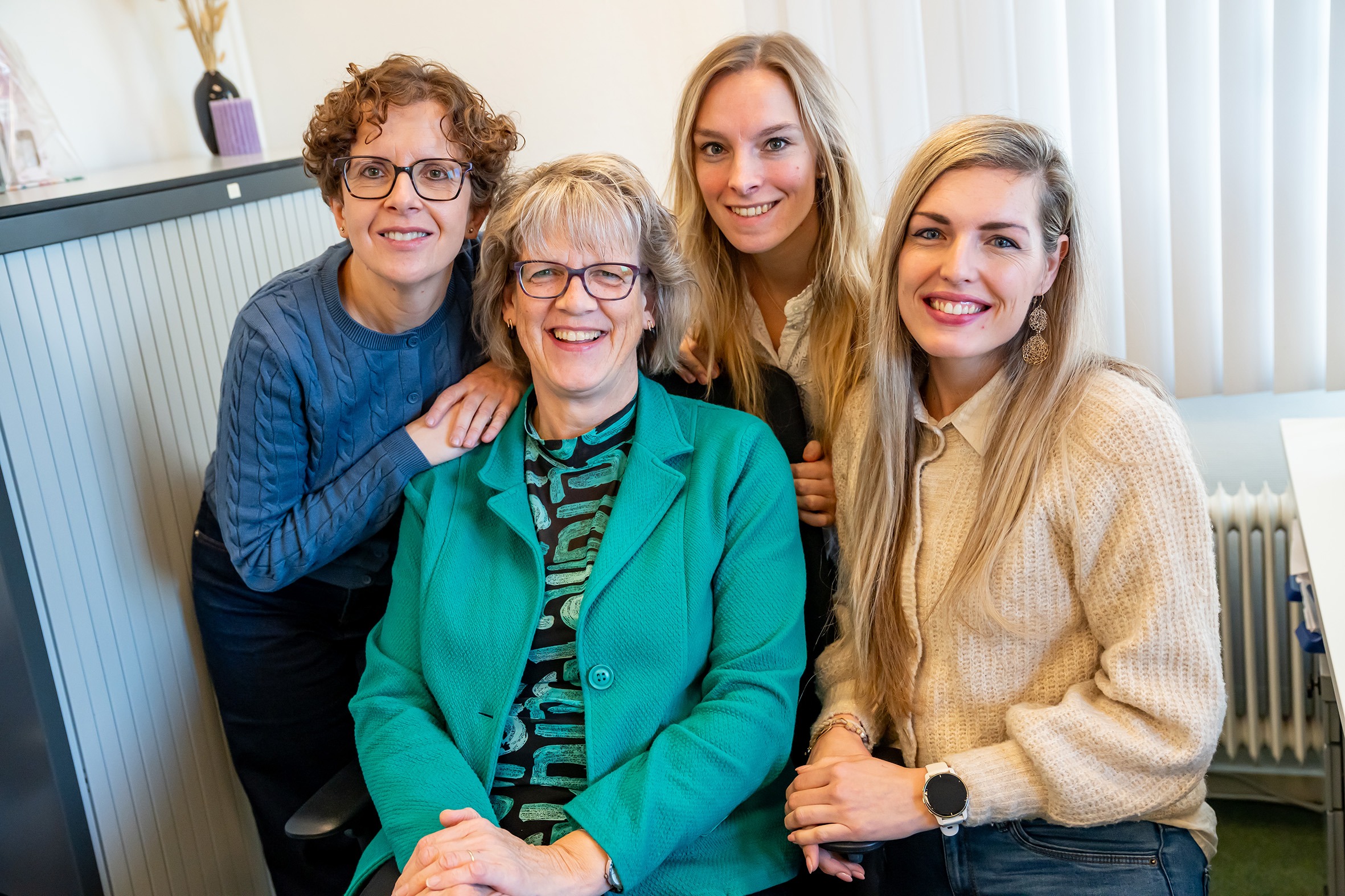 Teamfoto met vier lachende vrouwen op kantoor; één zit in een groen jasje, drie staan naast haar.