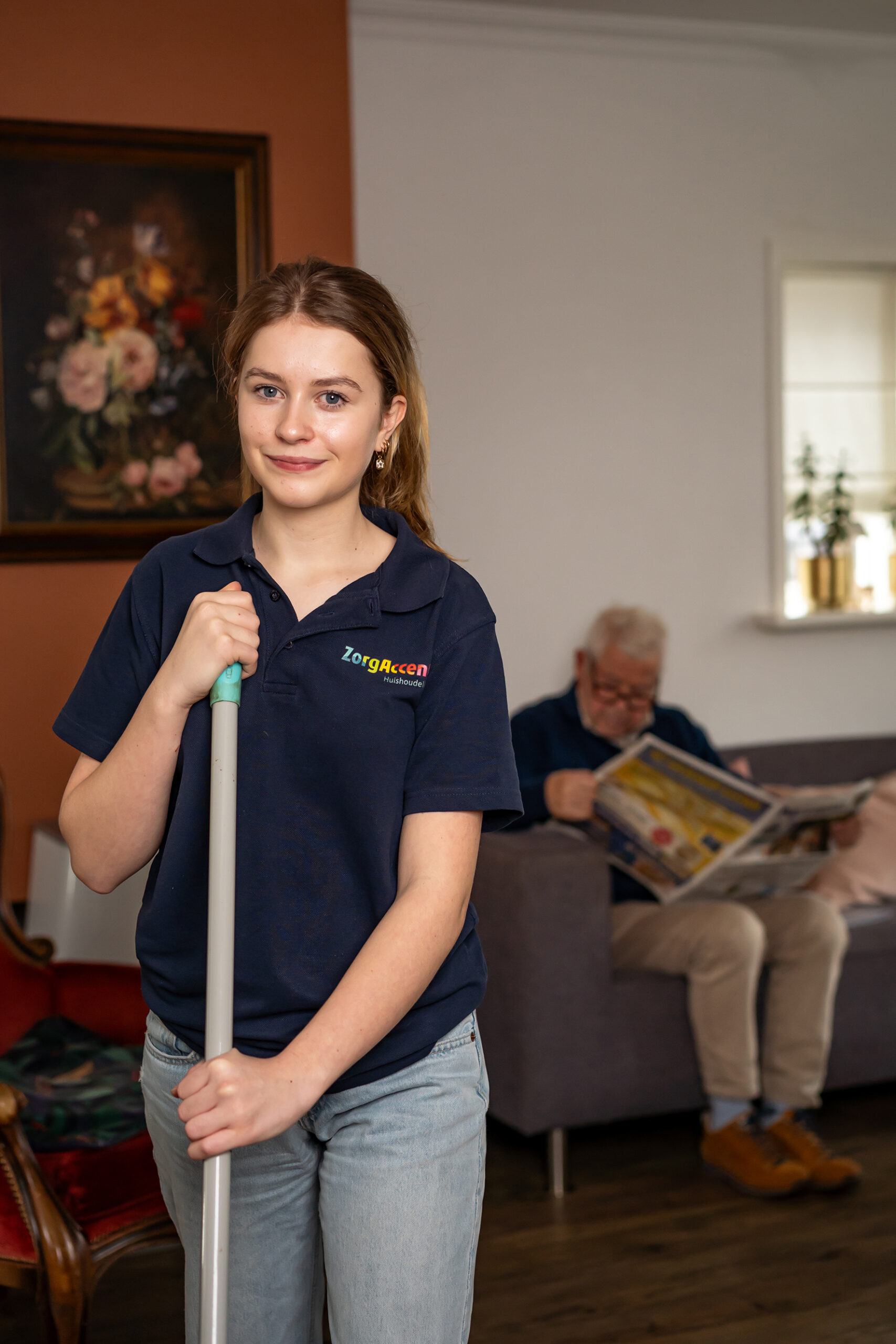 Huishoudelijke hulp met schoonmaaksteel in woonkamer, terwijl een oudere man op de bank de krant leest.