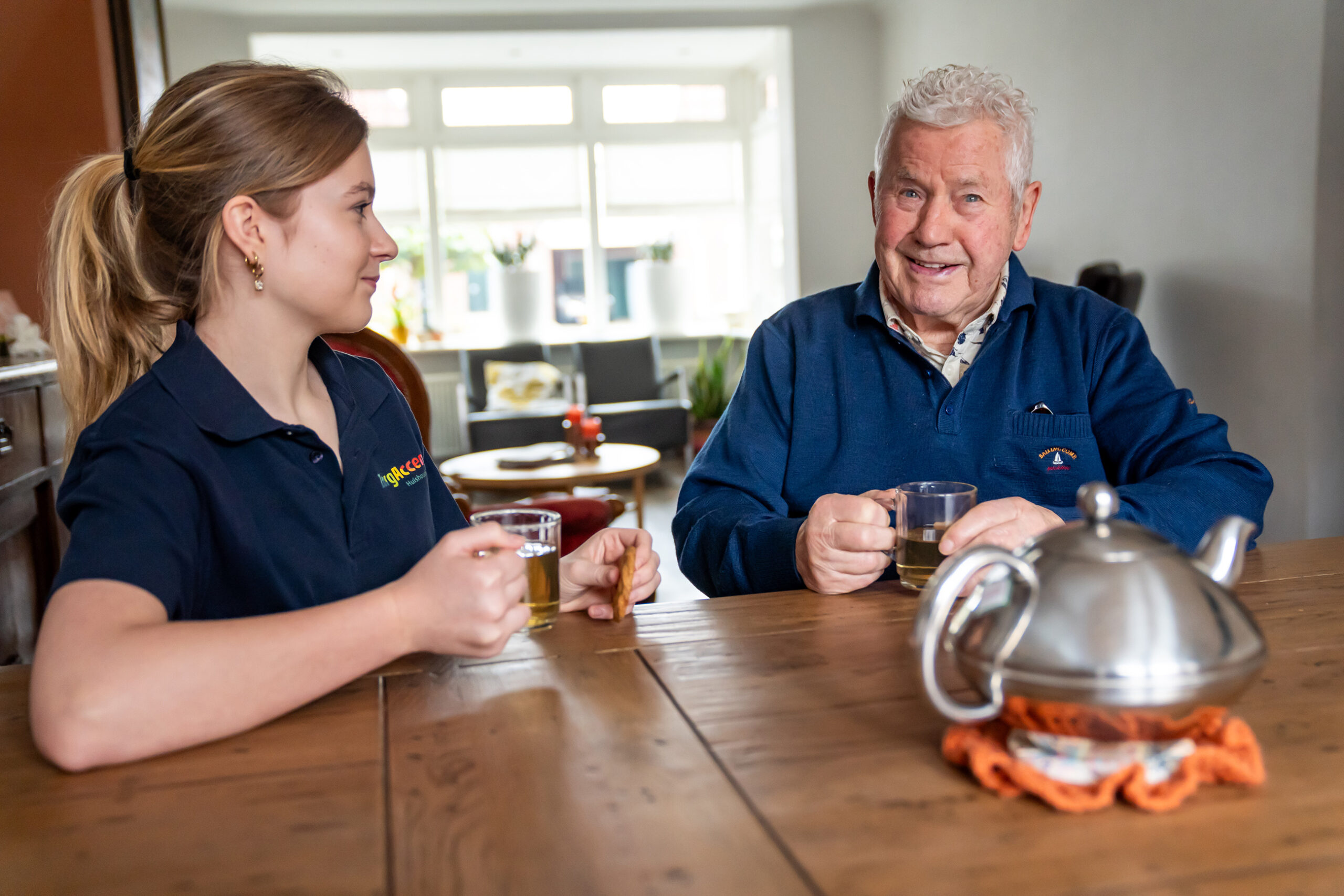 Jonge zorgmedewerker drinkt thee met oudere man aan de houten tafel in een lichte woonkamer.