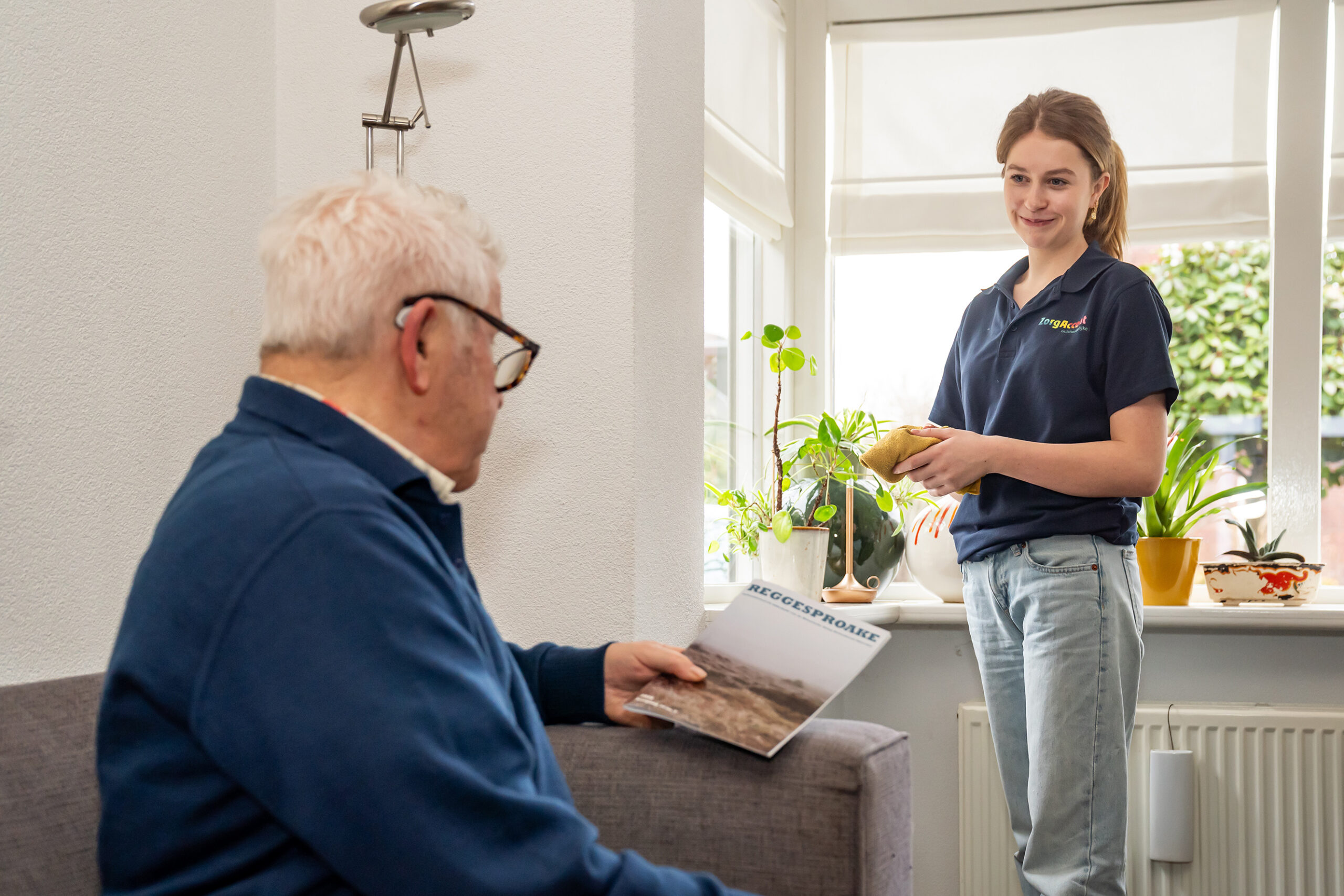 Oudere man met bril en gehoorapparaat leest, terwijl thuiszorgmedewerkster glimlachend stof afneemt bij het raam.