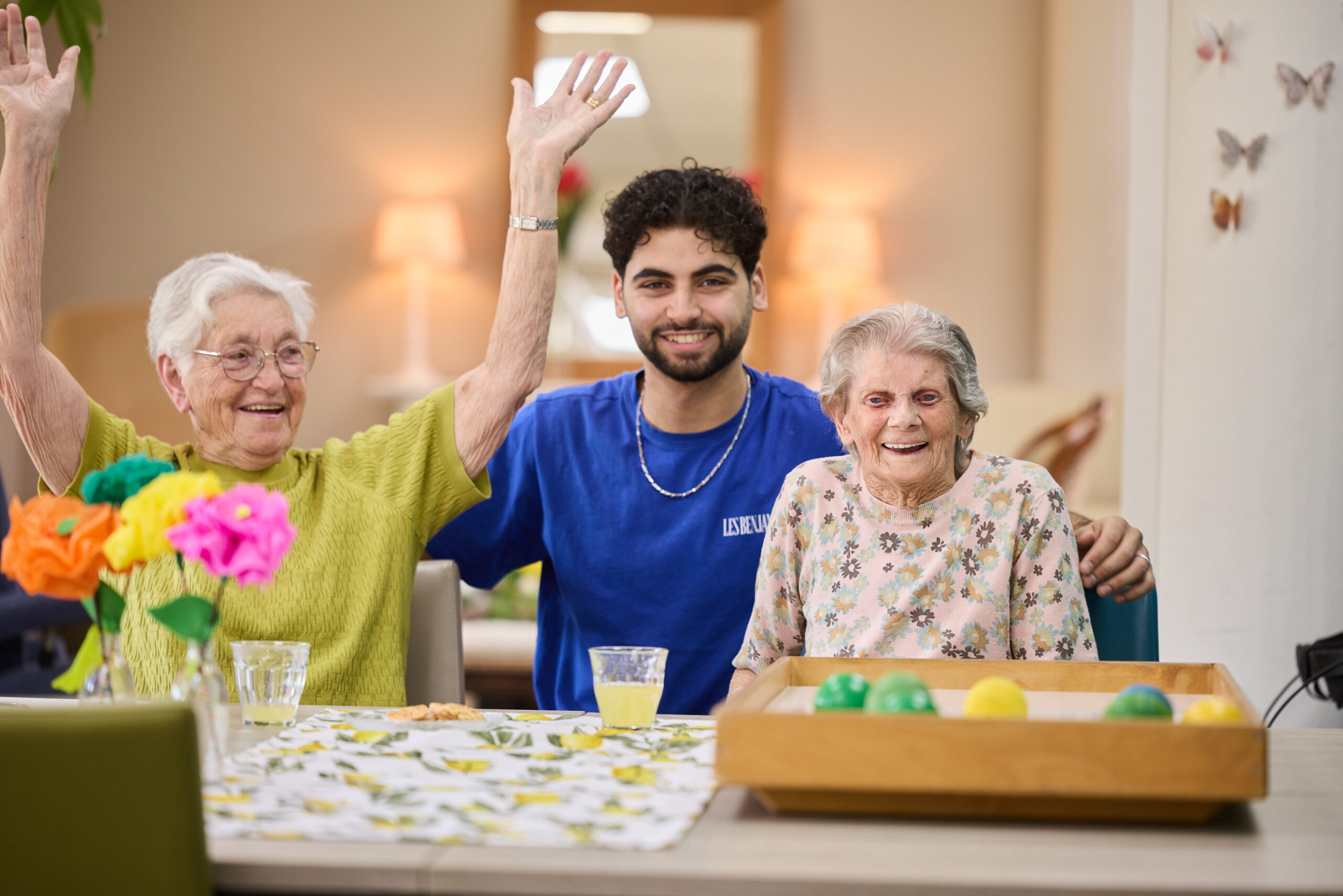 Jongeman met twee lachende oudere vrouwen aan tafel; één steekt juichend armen omhoog, spel met gekleurde ballen.