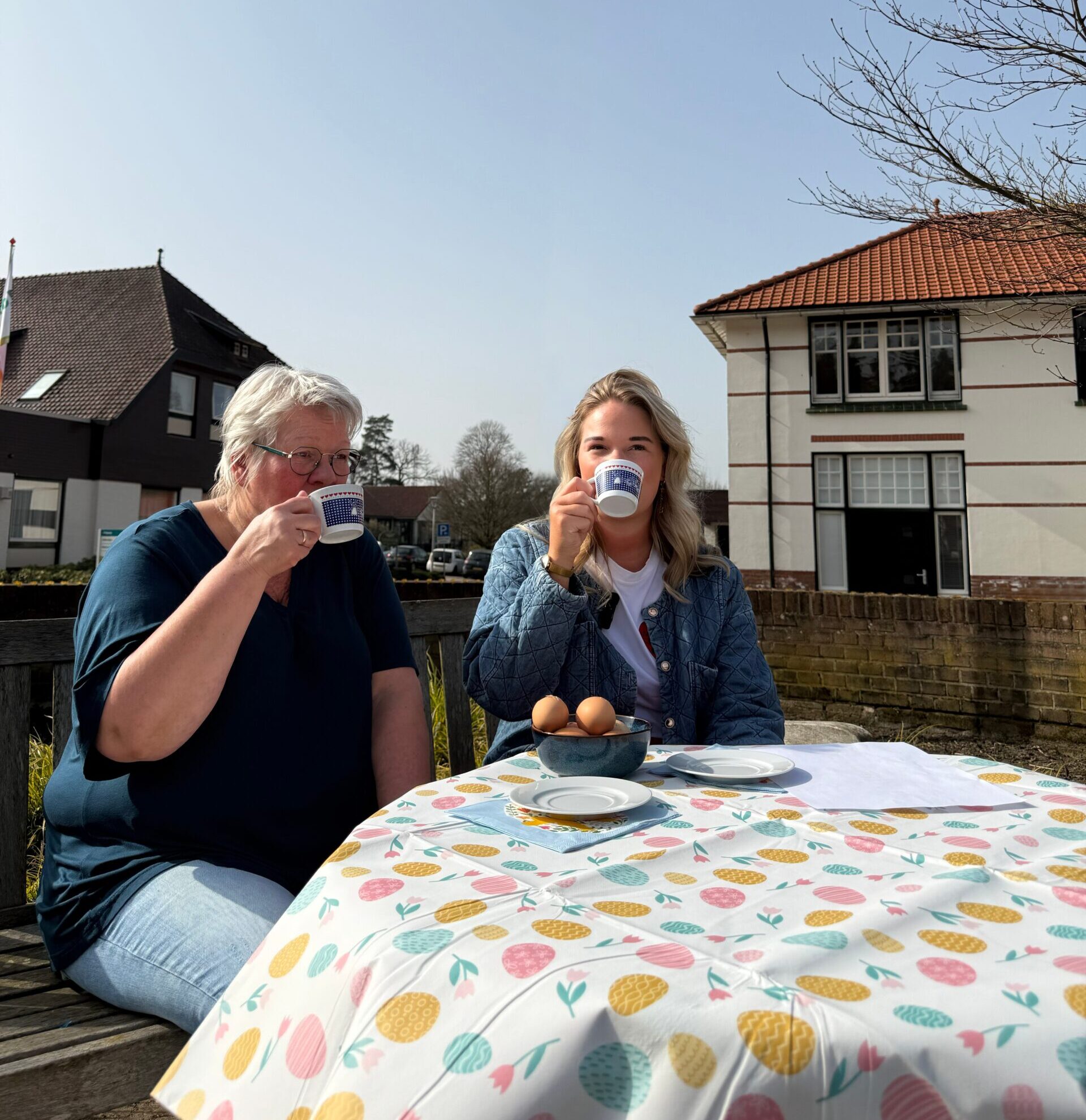 Twee vrouwen drinken koffie buiten aan een tafel met eieren, voor huizen op een zonnige lentedag.
