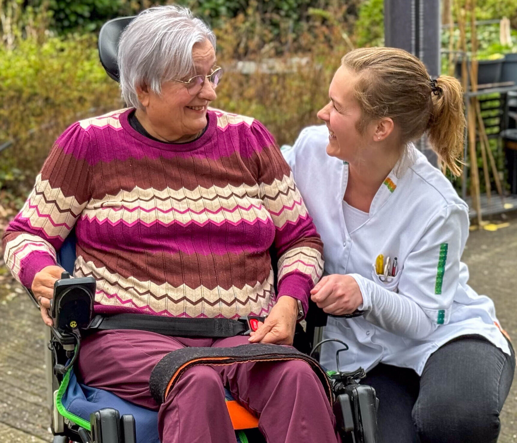 Oudere vrouw in rolstoel lacht met zorgverlener buiten in de tuin.