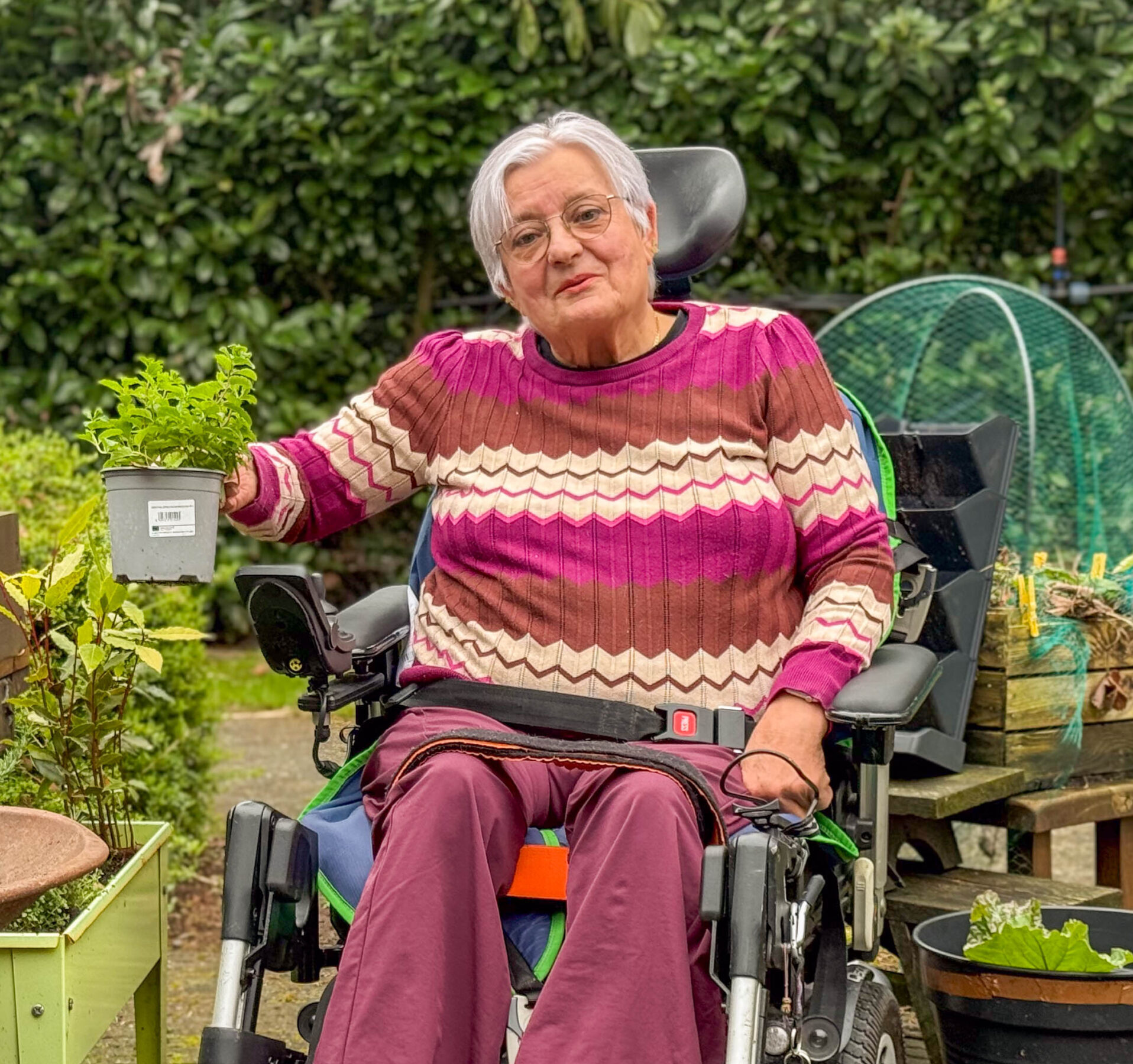 Vrouw in elektrische rolstoel houdt een potje met groen plantje vast in een tuin.