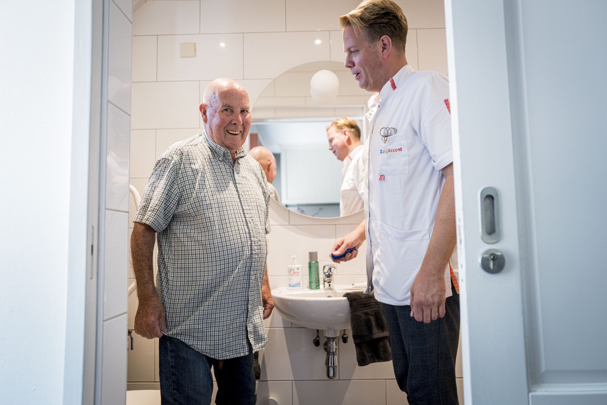 Verzorger in witte jas helpt oudere man in lichte badkamer bij tandenpoetsen; spiegel op de achtergrond.