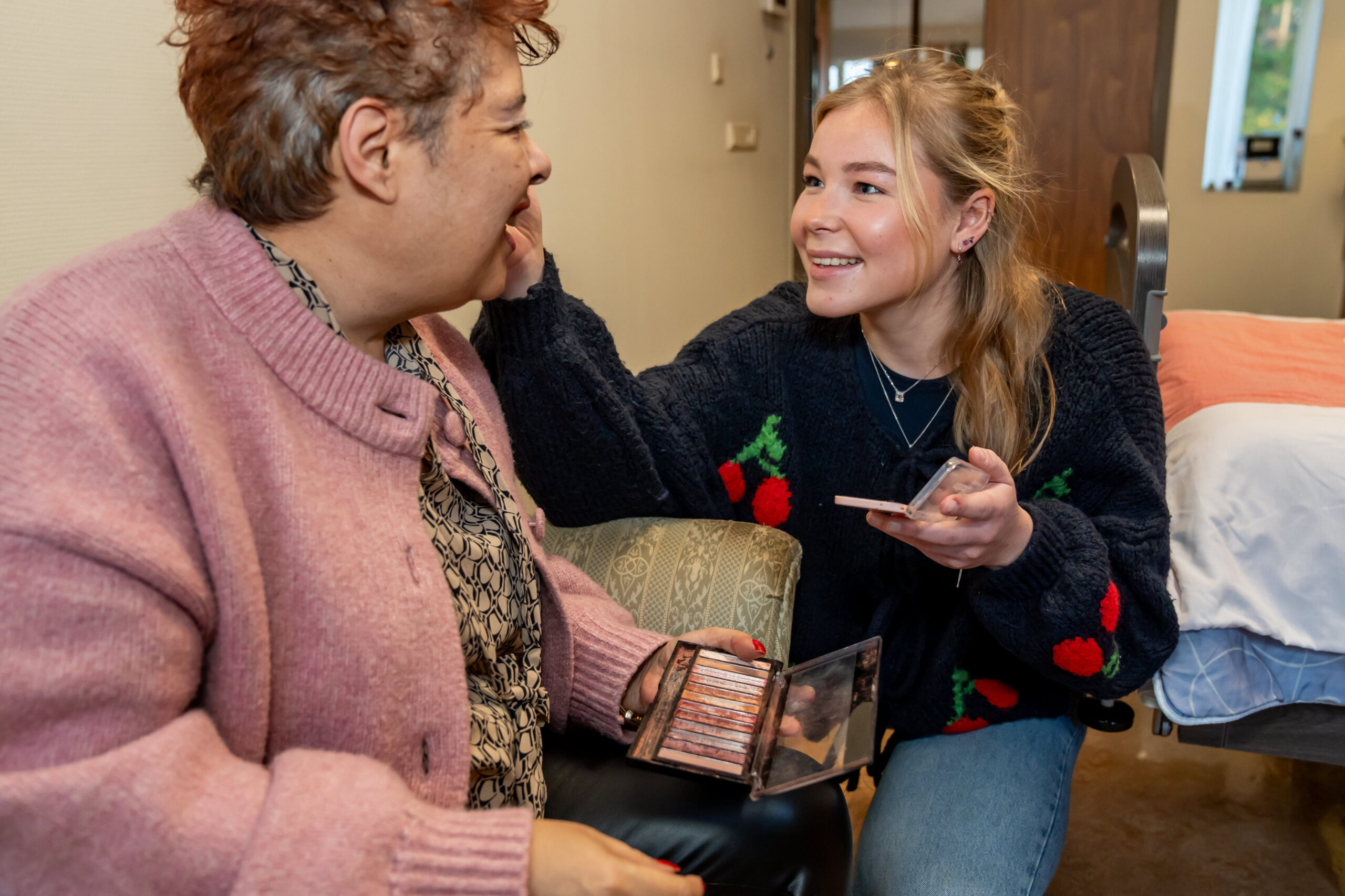 Jonge vrouw brengt lachend make-up aan bij een vrouw, met oogschaduwpalet in een kamer.