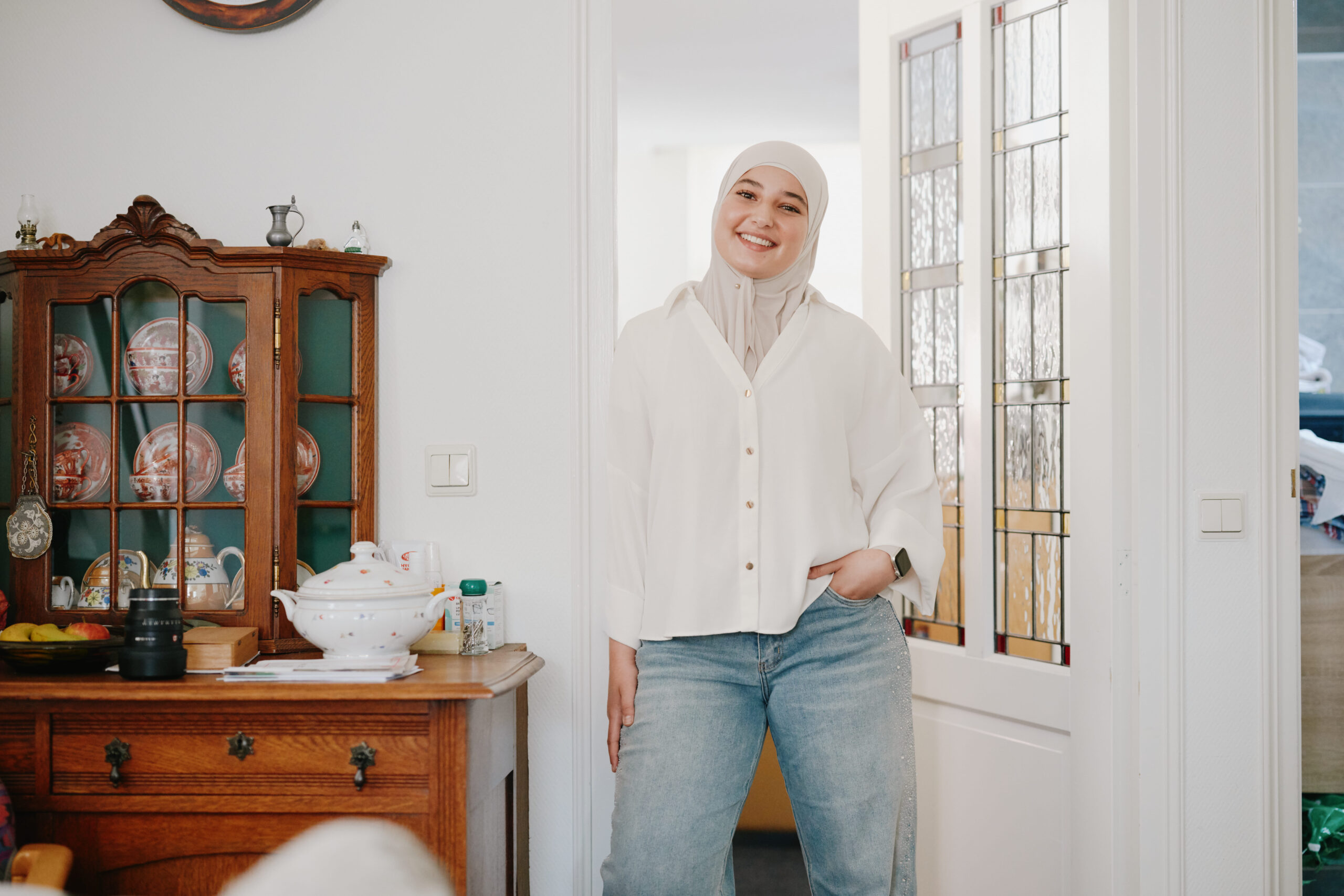 Lachende persoon met hoofddoek, witte blouse en spijkerbroek, staand in een gezellige woonkamer.