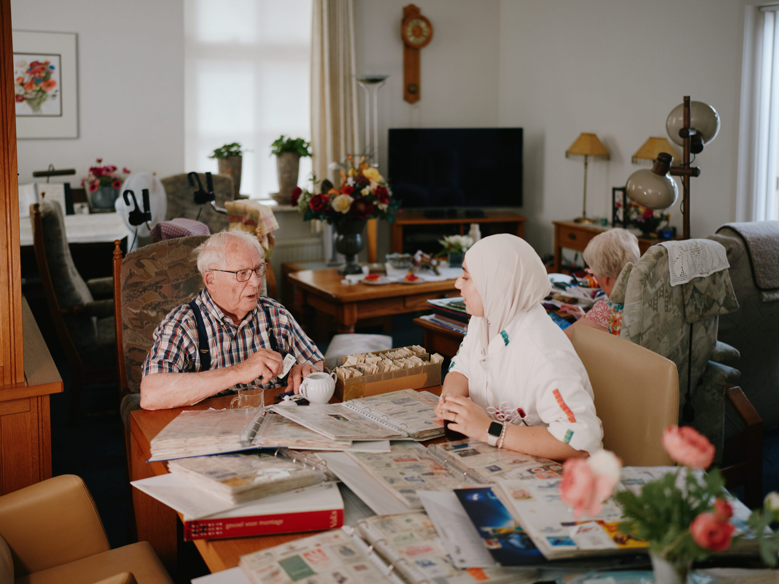 Oudere man en jonge vrouw met hoofddoek bekijken postzegelalbums aan een tafel in een gezellige woonkamer.